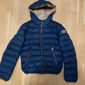 Blå dunjacka från Colmar - Snygg blå dunjacka från Colmar med huva och dragkedja. Jackan har ett stilrent märke på ärmen och är perfekt för kyligare dagar. Den quiltade designen ger både värme och stil.         Mått: längd = 51cm bredd 41,5cm 