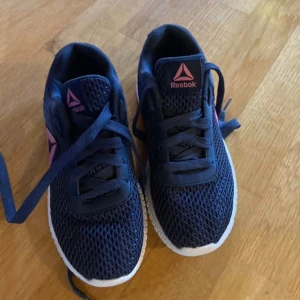 Mörkblå sneakers från Reebok - Snygga lätta mörkblå Reebok sneakers med rosa detaljer och snörning. Perfekta för en sportig look med en bekväm passform och stilren design. Lämpliga även för inomhussport.