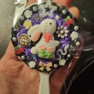 Mini handspegel  - Säljer en söt handspegel dekorerad med en vit kanin och blommor på en krämlim med lila och svart. Perfekt som en unik accessoar eller present. Storlek 10cm långt 5cm Bredd 