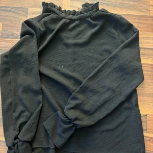 Svart blus från Boohoo - Säljer en elegant svart blus från Boohoo med volangdetaljer vid halsen. Blusen har långa ärmar och en knappstängning i nacken. Perfekt för en stilren look.