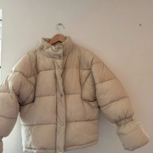 Beige dunjacka från NA-KD - Säljer en snygg beige dunjacka från NA-KD. Jackan har en oversized passform med långa ärmar och knappar framtill. Perfekt för kyliga dagar.