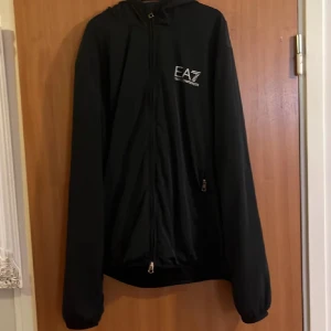 Svart jacka från Emporio Armani - Säljer en stilren svart jacka från Emporio Armani EA7. Jackan har en dragkedja framtill och en huva. Perfekt för en sportig look eller en avslappnad dag ute.
