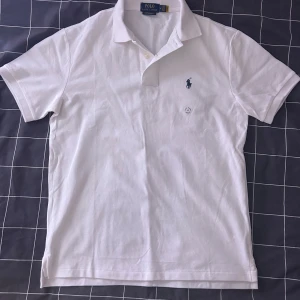 Vit kortärmad pikétröja från Ralph Lauren - Säljer en klassisk vit pikétröja från Ralph Lauren med korta ärmar och en broderad logga på bröstet. Perfekt för en stilren och avslappnad look. Tröjan har en krage med knappar.