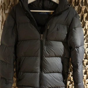 Grå dunjacka från Ralph Lauren - Säljer en snygg grå dunjacka från Ralph Lauren. Jackan har en quiltad design med dragkedja och en broderad logga på bröstet. Perfekt för kyliga dagar med sin varma och bekväma passform. Storlek XS men sitter mer som en S eller M.