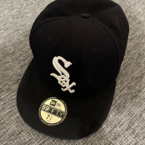 Svart keps från New Era - Snygg svart keps från New Era med broderad vit logotyp framtill. Modellen är 59FIFTY och har en platt skärm. Perfekt för en stilren look.