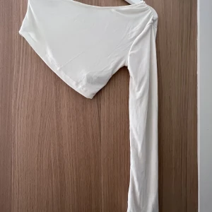 Oneshoulder crop top från NAKD - Aldrig använd!   Vit magtröja i väldigt fint och skimrigt material (se närbild). Armen är extra lång och något utsvängd. Mycket elegant! 