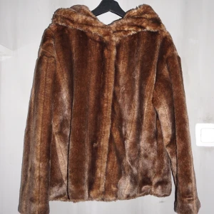 Brun Jacka i fuskpäls - Säljer denna pärla i brun fuskpäls som är perfekt till vardags och till kalla festtillfällen. Den har en stor fluffig luva som gör sig gott i snöfall. Den är i gott skick och hur skön som helst.