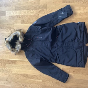 Oanvänd Jacka Vemdalen från märket Cobolt stl S/38 - Svart jacka från märket Cobolt.Vind- och vattentåligt yttermaterial, tre stycken rymliga fickor med dragkejdeförslutning. Den har muddar och kardborreband i ärmslut, samt luva med en dragsko och en fuskpäls som är avtagbar.  Helt oanvänd. Storlek S/38.