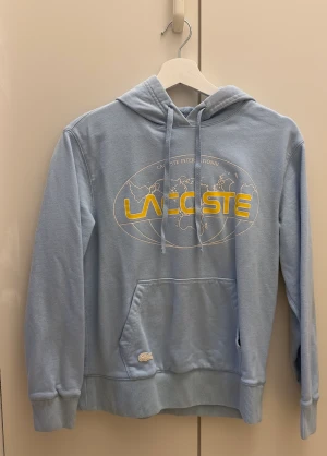 Ljusblå hoodie från Lacoste - En ljusblå hoodie med gula detaljer från Lacoste, sitter mer som en XS men är strl. S. Hoodien är använd i gott skick, MEN finns en defekt på hoodiens nedre mudd fram (se bild 3). Den blev missfärgad eller något när vi hade egen brunn, går med största san