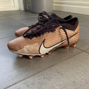 Nike Air Zoom Mercurial Vapor 15 Elite - Säljer ett par Nike Air Zoom Mercurial Vapor 15 Elite AG PRO Generation - guldbrun i storlek 39. Googla gärna och läs mer om detaljer på t ex unisport. Skorna är använda en säsong och säljer då de blivit för små. Slitage på undersidan enligt bild.