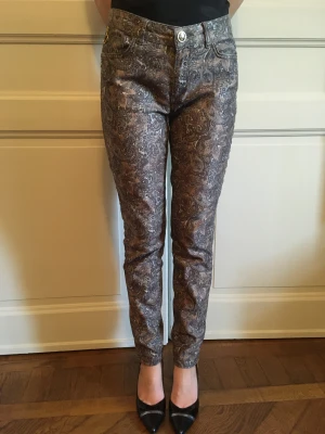 MOS MOSH byxor  - Mos Mosh Victoria Jive Pant - Metallic Choco Chip mönstrade byxor/jeans. Två framfickor, två bakfickor, dragkedja nere vid vristen, gulddetaljer. Midja är mellanväg.  Storlek: 27 Längd: 97 cm 