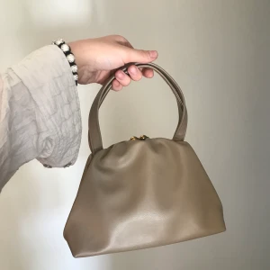 Väska - Säljer denna superfina beiga handväska som jag fyndat på second hand, men som tyvärr aldrig kommit till användning💕 perfekt storlek för att få med sig det nödvändiga! Superfint knäppe i guld som ger väskan det lilla extra! Skriv om ni vill ha fler bilder💓