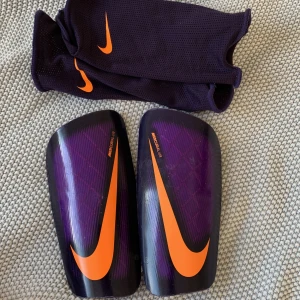 Benskydd Nike  - Snygga benskydd från Nike mercurial/lite. Använda men i bra skick. Helt nytvättade i maskin. Lila med svart ytterkant och neonorangea detaljer. Fotbollsskor finns i annat inlägg