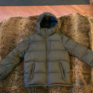 Grå Polo Ralph Lauren Jacka - Säljer en Ralph Lauren Jacka eftersom det blivit förliten väldigt snabbt. Köpte den förra vintern för ca 2500kr. Den är 9/10 skick eftersom den har en liten skråma ovanför märket skriv om bilder. Priset kan diskuteras.