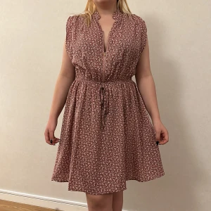 Sommarklänning  - Ljusrosa blommig klänning. Dragkedja så man kan hur v-ringad man vill ha det. 