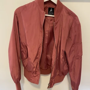 Rosa bomberjacka - Metallrosa färg. Jackan har dragkedja och fickor framtill och på ärmen. Elastisk slutning på ärmar och vid midjan. 