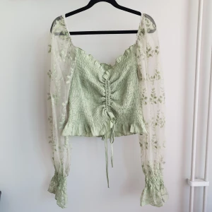Topp - Grön långärmad topp med ärmar i mesh med ett snyggt broderat mönster. Något ceoppad modell med knytning framtill för att kunna reglera längden. Är väldigt strechig. Har aldrig använts utan är bara testad.