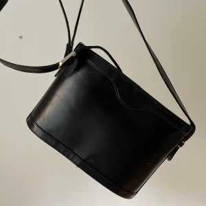 Skinnväska  - Oval axelremsväska i svart skinn med dragkedja och detaljer i guld.