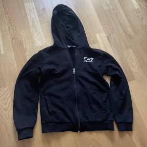 Svart zip up hoodie från märket EA7. Skicket på tröjan 8/10. Storlek: 152.