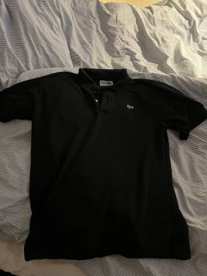 Lacoste Piké  - Svart lacoste piké i tunt löst material.  Inga flaws som hål eller missfärgning. Storlek medium i tagen men skulle säga att den är mer som s/m