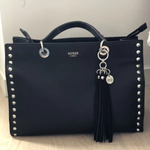 GUESS VÄSKA  - Guess väska   Svart utsida, beige tyg på insidan med ljus bruna guess märkningar.   Silver detaljer och dragkedja.    4 innefickor.  Finns ett avtagbart långt band till. (”Tassel” även avtagbar)  Helt oanvänd.   
