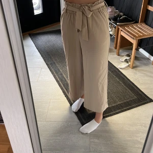 Kostymbyxor  - Beigea vida kostymbyxor med risår i midjan som täcks av en rosett. 