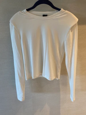 Tröja  - Vit tröja med axel vaddering. Armlängd på 50 cm och längd på 30 cm från armhålan. Inte annvänd. 
