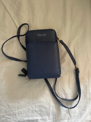 Marinblå handväska - Denna smidiga väskan är aldrig använd och är i fint skick. Finns många smarta fack med plats för mobiltelefon, nycklar mm. Finns även innyggd korthållare. Kommer även med kortare band så man kan ha den endast runt handleden om man vill. Perfekt till resor