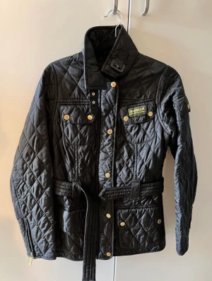 Barbour International quilt jacket  - Quiltad vårjacka med fyra fickor fram och bälte i midjan samt en liten ficka på ärmen. Barbour International logotyp på vänster bröst. 