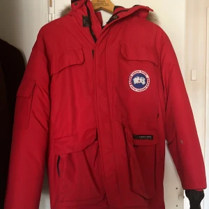 Röd Canada Goose jacka  - Säljer min röda Canada Goose jacka då jag ej använder den och tänker att denna jacka ska värma någon annan.  Jackan är i gott skick men tecken på användning finns. Den har inga  fläckar, hål eller sådana skråmor.  Dragskedjan är lite småkrånglig. 