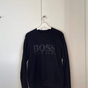 Svart Hugo boss sweatshirt med vit, svart, grön och blå text. Tröjan är använd men i bra skick. Storlek M