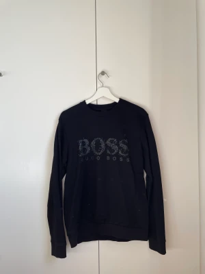 Hugo boss tröja - Svart Hugo boss sweatshirt med vit, svart, grön och blå text. Tröjan är använd men i bra skick. Storlek M