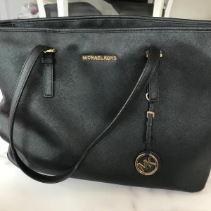 Äkta Michael kors väska - Svart Michael kors väska, orginalmodellen skulle jag säga, har tex ett fack där man kan lägga sin dator och andra smarta fack i väskan. Har lite defekter så be om ber bilder om ni är intresserade, men defekterna syns inte tydligt. Köpt för 3000kr