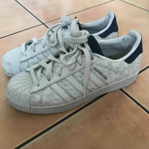  - Originella Adidas Superstar som är vita och blåa med ett diskret tryck på. Innersulan är lite smutsig men går enkelt att byta ut. Blåa detaljer där bak som blir som reflexer med flash. Storlek 39 1/3 