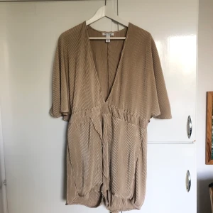 Shortsdress med band  - En byx/shortsdress från Nelly. Guld i färgen med tillhörande band för att knyta i midjan om så önskas. Storlek: XL. Frakt: 44 kr ej spårbart (63 kronor med postnord spårbart). 