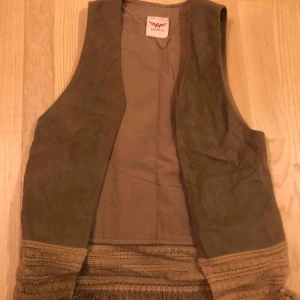  - Moccaväst i beige/naturfärg, strl S. Mocca på utsidan av västen. Västen har fransar längst ner. 