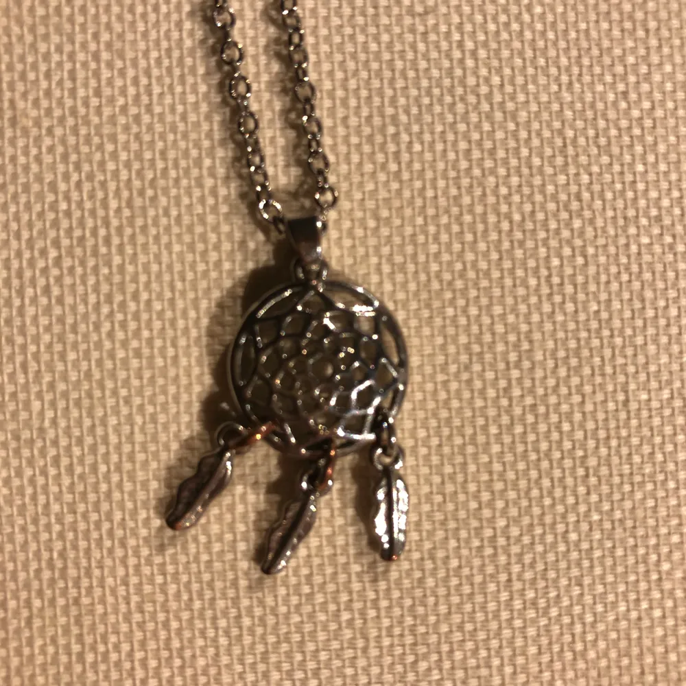 Ett silverfärgat halsband med en liten drömfångare som berlock. Asusteet.