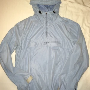  - Ljusblå pullover jacka med stor ficka på magen och skärm på luvan. Står som "female" M, känns som en S på mig. Funkar utmärkt som festivaljacka! FTP broderat på kanten utav fickan. 