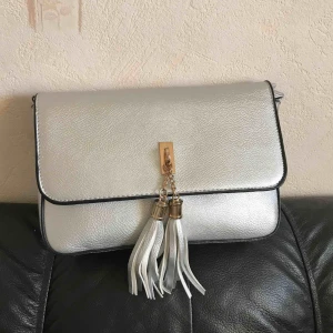 Silvrig corssbody väska med tassle  - L: 24cm B:17cm D:6cm köpt på Hollys butiken i Barkaby. Använt 2 gånger totalt. Jättefin silvrig väska med gulddetaljer och två tassels. Säljer pga att den inte kommer till  användning.