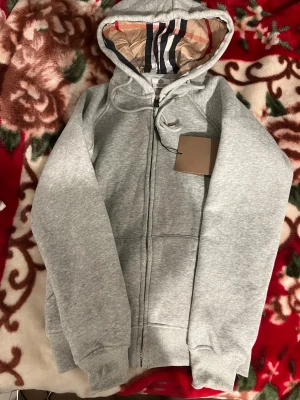 Grå zip hoodie från Burberry - Snygg grå hoodie från Burberry med dragkedja framtill och klassiskt rutigt foder i huvan. Hoodien har justerbara snören i huvan och ribbade muddar vid ärmar och nederkant. Perfekt för en chill och stilren look.