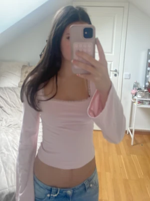 Ljusrosa långärmad topp med volang - Söt ljusrosa långärmad topp med volangdetaljer vid ärmslut och en subtil spetskant vid halsen. Toppens passform är tight och den är croppad, perfekt att matcha med jeans. Materialet ser ut att vara bomull eller polyester.