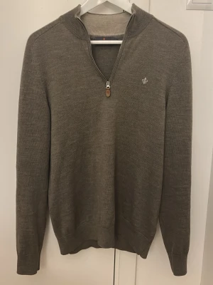 Merinowoll brun stickad tröja med halvzip (Morris) - Snygg brun stickad tröja med halvzip och hög krage. Tröjan har ett diskret broderat emblem på bröstet och ribbade muddar vid ärmslut och nederkant. Perfekt för lager på lager och en clean look.