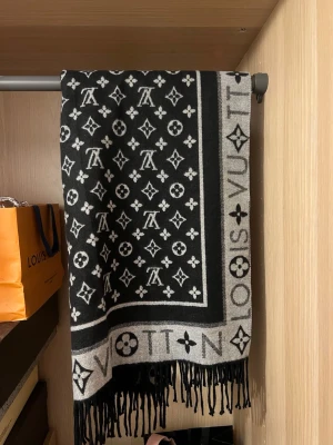 Louis Vuitton svart och grå halsduk - Snygg halsduk från Louis Vuitton i svart och grått med klassiskt monogrammönster och logotyp längs kanten. Halsduken har fransar i ändarna och är vändbar med olika färgkombinationer på varje sida. Perfekt accessoar för att lyfta din outfit.