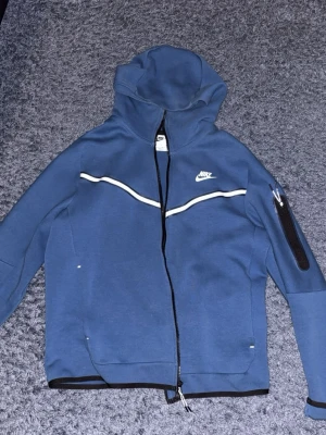 Blå Nike hoodie med dragkedja - Snygg blå hoodie från Nike med hel dragkedja och vit logga på bröstet. Hoodien har en huva, vita detaljer över bröstet och en praktisk ficka med dragkedja på ena ärmen. Perfekt för en sportig och avslappnad stil.