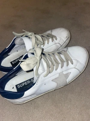 Golden Goose vita sneakers med stjärna - Kom med prisförslag, Golden Goose sneakers i vitt läder med grå mockastjärna på sidan och mörkblå häl. Beige skosnören och låg profil. Klassisk Super-Star modell med retrovibe och coola detaljer. Perfekta för dig som gillar streetstyle och vill ha något unikt.