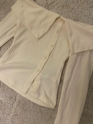 Offshoulder ribbad topp med knappar - Trendig offshoulder topp i ljus beige ribbad bomullsmix med långa ärmar och knappar framtill. Snygg bred krage som ger en unik look. Perfekt för dig som vill sticka ut lite extra.