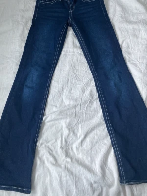 Mörkblå bootcut jeans med kontrastsömmar - Snygga mörkblå bootcut jeans med vita kontrastsömmar och klassisk femficksdesign. Jeansen har låg midja och är tillverkade i stretchigt denimtyg för en bekväm passform. Perfekta för dig som gillar en tidlös och cool look.