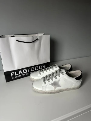 Golden Goose vita  - Snygga vita sneakers från Golden Goose med grå stjärna på sidan, grå skosnören och svart detalj på hälen. Skorna har en klassisk låg siluett och är tillverkade i läder med slitna detaljer för en cool, avslappnad look.