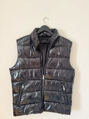 Svart glansig dunväst från Righteous - Snygg svart dunväst från Righteous med glansig finish, nästan helt oanvänd. Västen har två dragkedje fickor och en diskret logga på bröstet.            Pris är inte hugget i sten!