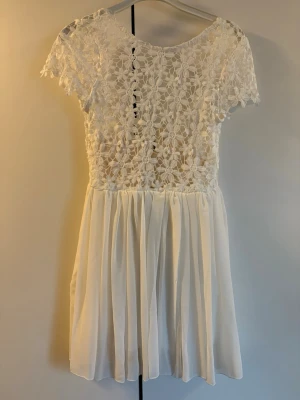 Vit spetsklänning med kort ärm - Söt vit kortklänning med överdel i blommig spets och kjol i lätt, flödande tyg. Klänningen har korta ärmar och rund halsringning. Perfekt för sommarens alla tillfällen och ger en romantisk vibe.